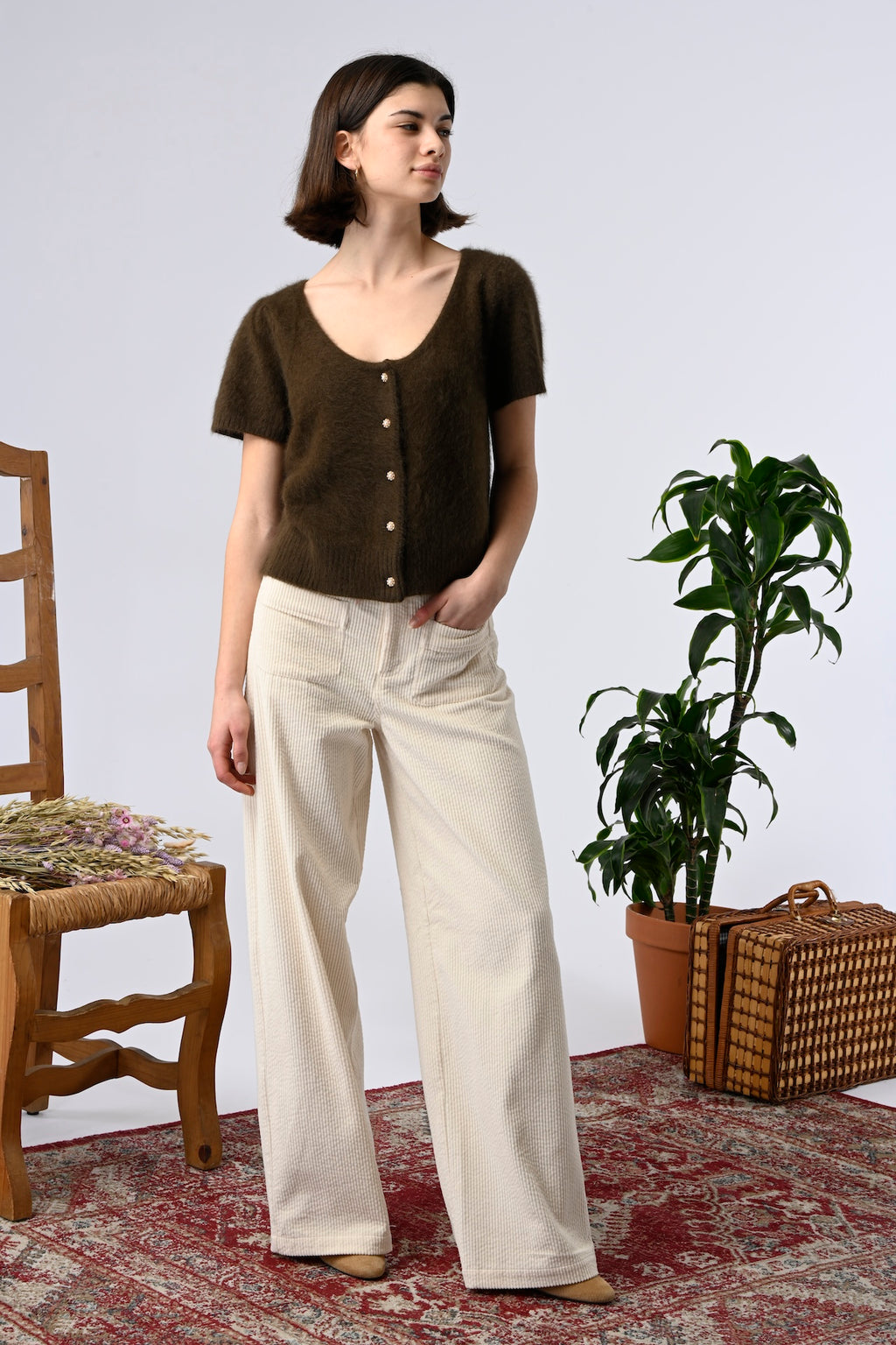 Woman wearing a brown top with erru patch pocket pants standing in a room with a wooden chair, plant, and basket.