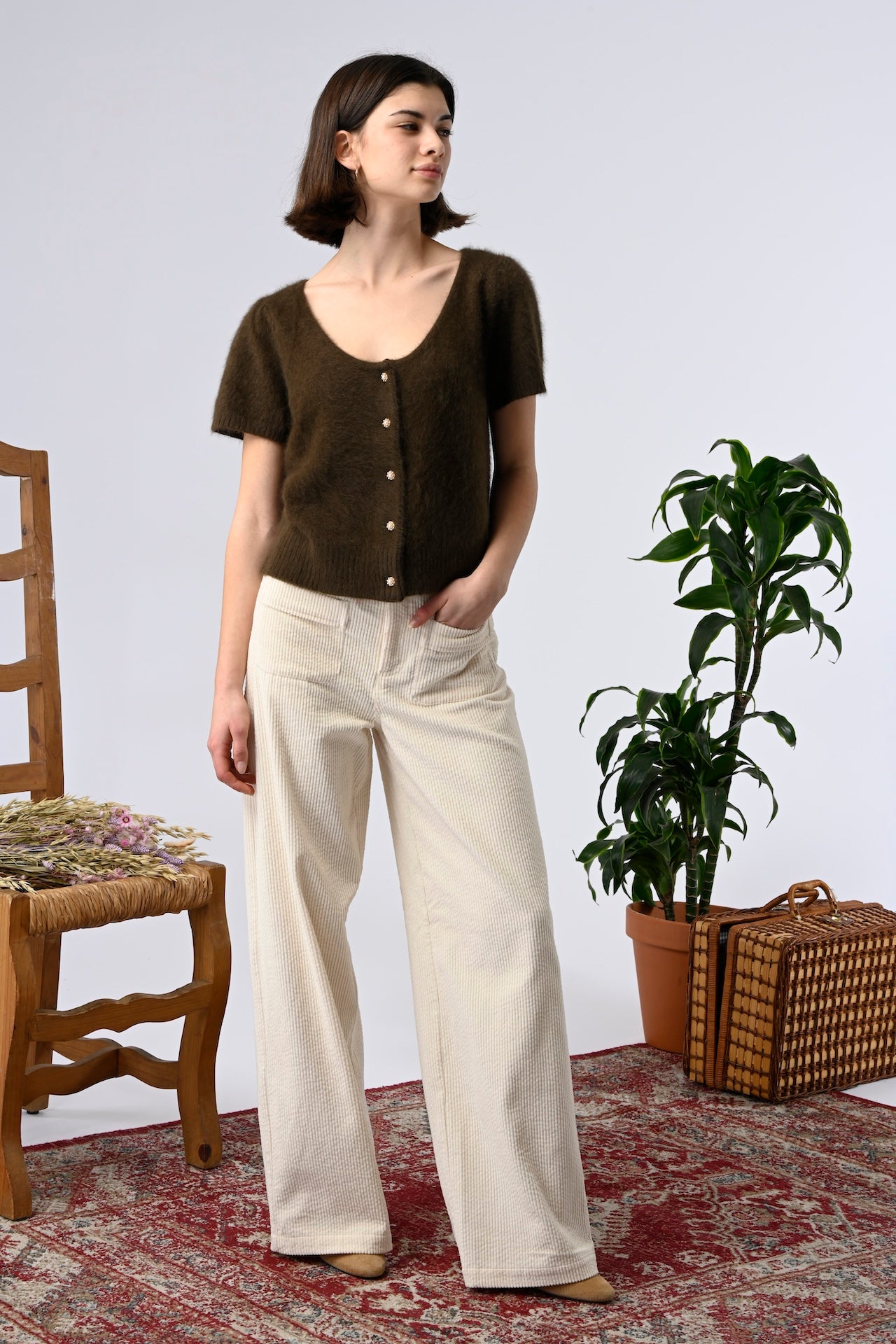 Woman wearing a brown top with erru patch pocket pants standing in a room with a wooden chair, plant, and basket.