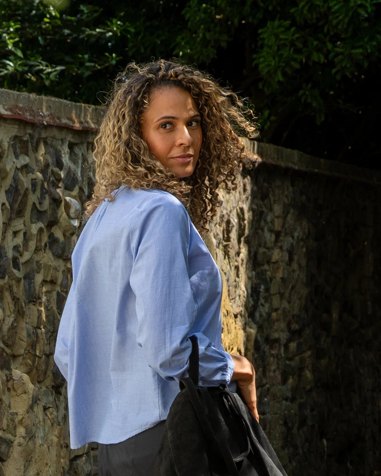 Woman in a light blue Chalk shirt with pearl button standing against a stone wall with greenery.