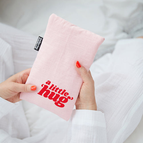 Pink pillow with 'a little hug' text held by a person in a white robe.